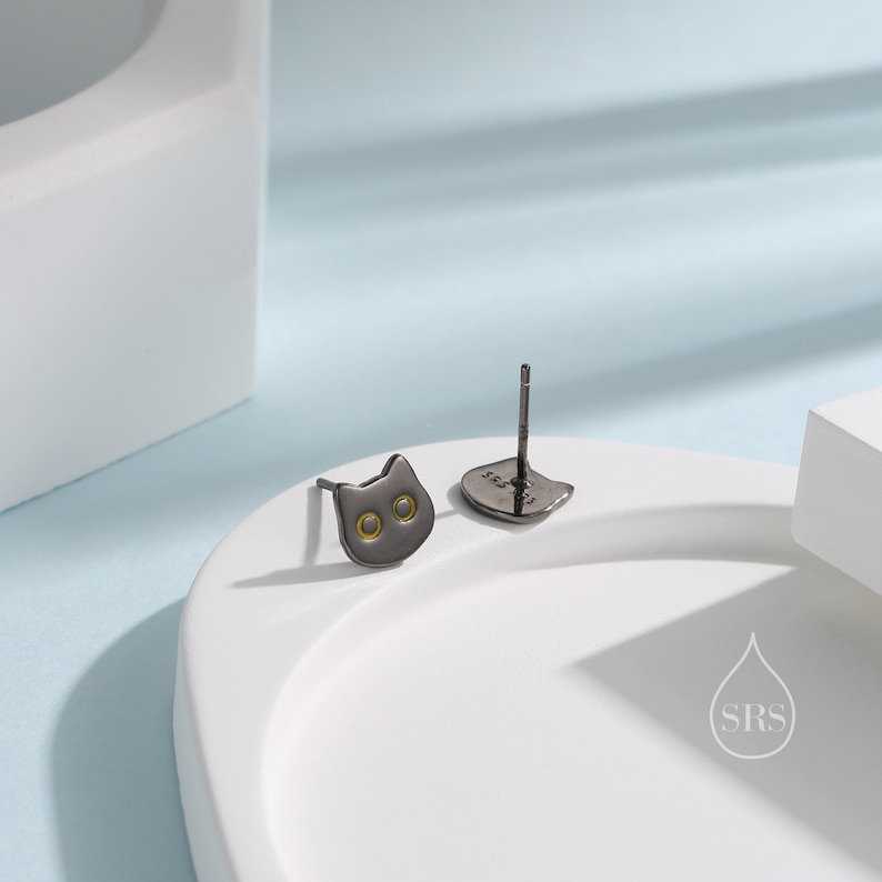 Peut inclure: Une paire de boucles d'oreilles clous en forme de chat noir avec des accents dor&eacute;s. Les boucles d'oreilles sont sur une surface blanche.