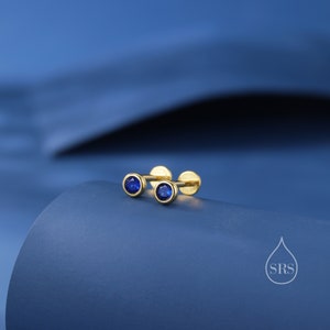 Puede incluir: Un par de pendientes de botón de oro con pequeñas piedras azules. Los pendientes están sobre un fondo azul.