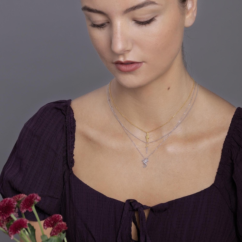 May include: A layered necklace set consisting of three delicate chains. The top chain is gold-toned with a lightning bolt pendant. The middle chain is silver-toned with a small cross pendant. The bottom chain is silver-toned with a dragon pendant.