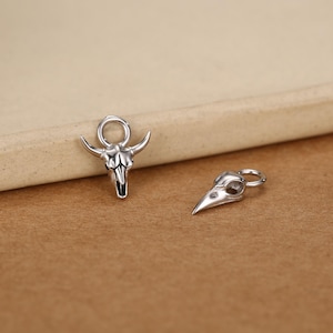 May include: Two silver-toned metal charms featuring detailed animal skull designs. One charm depicts a bull skull with prominent horns, while the other charm represents a bird skull with a pointed beak. Both charms include a small circular jump ring.