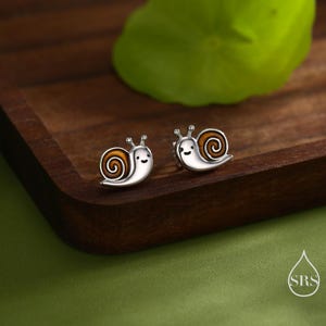 Può includere: Un paio di orecchini a lobo a forma di lumaca in argento, con dettagli in smalto marrone e bianco. Le lumache hanno facce sorridenti e sono appoggiate su un vassoio di legno scuro. Un accessorio carino e stravagante.