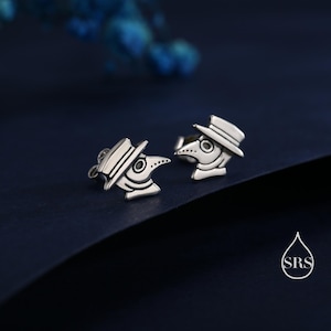 May include: A pair of silver-colored stud earrings. Each earring is shaped like a plague doctor's head, complete with a hat and beak. The earrings are set against a dark blue background, with a small logo in the bottom right corner.