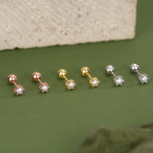 May include: An assortment of stud earrings in rose gold, gold, and silver tones. Each earring features a small, round pearl at the center of a decorative setting. The earrings are displayed on a green surface, with a neutral-colored stone in the background.