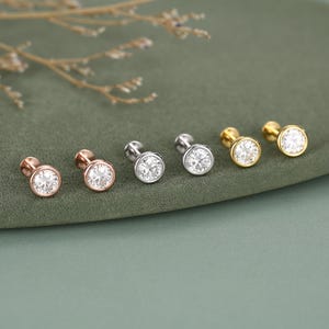 May include: Five stud earrings in rose gold, silver, and gold. Each earring has a round, clear gemstone in a bezel setting. The earrings are displayed on a green surface with a sprig of dried flowers. The earrings are suitable for pierced ears.