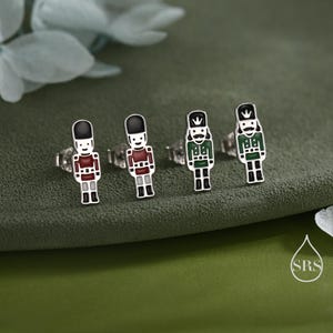 May include: Four pairs of silver-toned stud earrings. Each earring features a colorful, miniature soldier design with black, red, and green accents. The earrings are set against a green background.