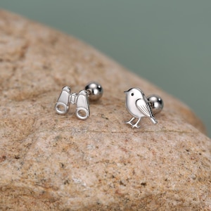 May include: A pair of silver-toned stud earrings featuring a bird-shaped design on one earring and a pair of binoculars on the other. Each earring has a small spherical backing and a polished metallic finish, showcasing detailed engravings of the bird.