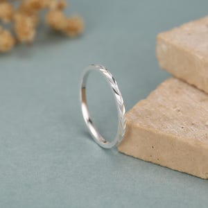 Può includere: Un sottile anello in argento caratterizzato da un motivo ripetitivo a chevron o a forma di foglia inciso lungo la fascia esterna. Il metallo presenta una finitura lucida e riflettente che cattura la luce, creando piccole sfaccettature angolari.