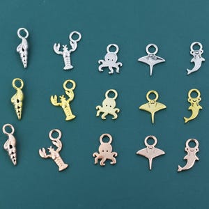 May include: A collection of silver, gold, and rose gold sea-themed charms. The charms include a seashell, lobster, octopus, stingray, and a fish. The charms are arranged on a teal background.