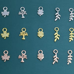 Könnte beinhalten: Eine Sammlung von Metallanhängern in verschiedenen Formen, darunter Ahornblätter, Bäume, Kleeblätter und Zweige. Die Anhänger sind in Silber-, Gold- und Roségoldtönen gehalten und auf einer dunkelgrünen Oberfläche angeordnet. Jeder Anhänger hat eine kleine Schlaufe.