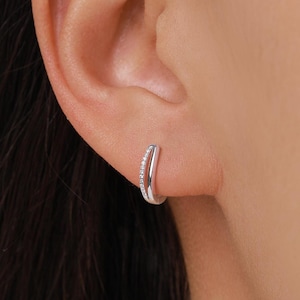 May include: A silver hoop earring with a row of small, clear gemstones along the outer edge. The earring is worn on an ear, showcasing its curved design and sparkling details. The jewelry is simple and elegant.