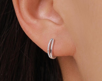 May include: A silver hoop earring with a row of small, clear gemstones along the outer edge. The earring is worn on an ear, showcasing its curved design and sparkling details. The jewelry is simple and elegant.