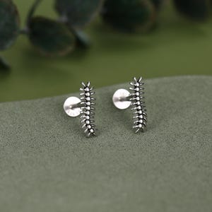 Könnte beinhalten: Ein Paar Ohrstecker aus Silber in Form von Tausendfüßlern. Jeder Ohrstecker hat einen langen, segmentierten Körper mit mehreren kleinen Beinen, die von beiden Seiten abstehen. Das Metall hat ein oxidiertes Finish, um die Textur hervorzuheben.