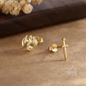 Peut inclure: Boucles d'oreilles dor&eacute;es. L'une repr&eacute;sente un dragon aux ailes d&eacute;ploy&eacute;es, l'autre une &eacute;p&eacute;e. Les boucles d'oreilles sont pr&eacute;sent&eacute;es sur une surface marron avec un arrangement floral flou en arri&egrave;re-plan.