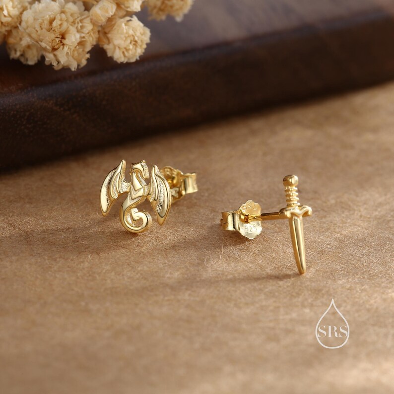 Peut inclure: Boucles d'oreilles dor&eacute;es. L'une repr&eacute;sente un dragon aux ailes d&eacute;ploy&eacute;es, l'autre une &eacute;p&eacute;e. Les boucles d'oreilles sont pr&eacute;sent&eacute;es sur une surface marron avec un arrangement floral flou en arri&egrave;re-plan.