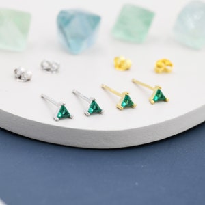 May include: A selection of small stud earrings featuring triangular, emerald-green gemstones. The earrings are presented in pairs, with silver and gold metal options. Earring backs are also visible. The background is a soft white with hints of blue and green.