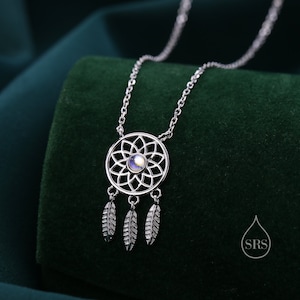 May include: A silver-toned necklace featuring a circular pendant with a symmetrical floral cutout design and a central iridescent stone. Three small, textured silver-toned feather charms hang from the bottom of the circular frame on a delicate chain.