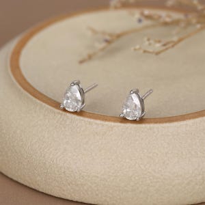May include: A pair of silver stud earrings with pear-shaped, clear gemstones. The earrings are set in a silver metal and are displayed on a beige surface. The earrings are a classic jewelry item.