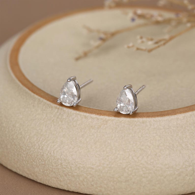 May include: A pair of silver stud earrings with pear-shaped, clear gemstones. The earrings are set in a silver metal and are displayed on a beige surface. The earrings are a classic jewelry item.