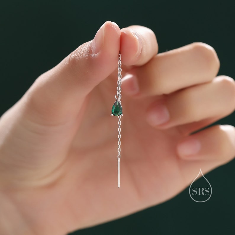 May include: A delicate silver earring featuring a teardrop-shaped, emerald-green gemstone. The earring hangs from a fine chain and a straight bar. The earring is held up by a hand against a dark green background.