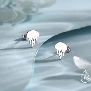 Puede incluir: Dos pendientes de plata con forma de medusa. Los pendientes tienen un acabado liso y pulido y están colocados sobre un fondo azul claro. El diseño de la medusa presenta una parte superior redondeada y tentáculos fluidos.