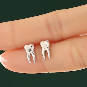 May include: A pair of small, polished silver-toned stud earrings shaped like human molars. Each earring features a smooth, metallic surface with a distinct two-rooted design, reflecting light across its curved, anatomical form.