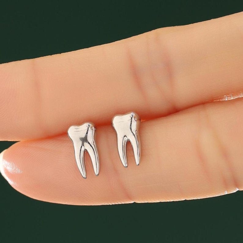 May include: A pair of small, polished silver-toned stud earrings shaped like human molars. Each earring features a smooth, metallic surface with a distinct two-rooted design, reflecting light across its curved, anatomical form.