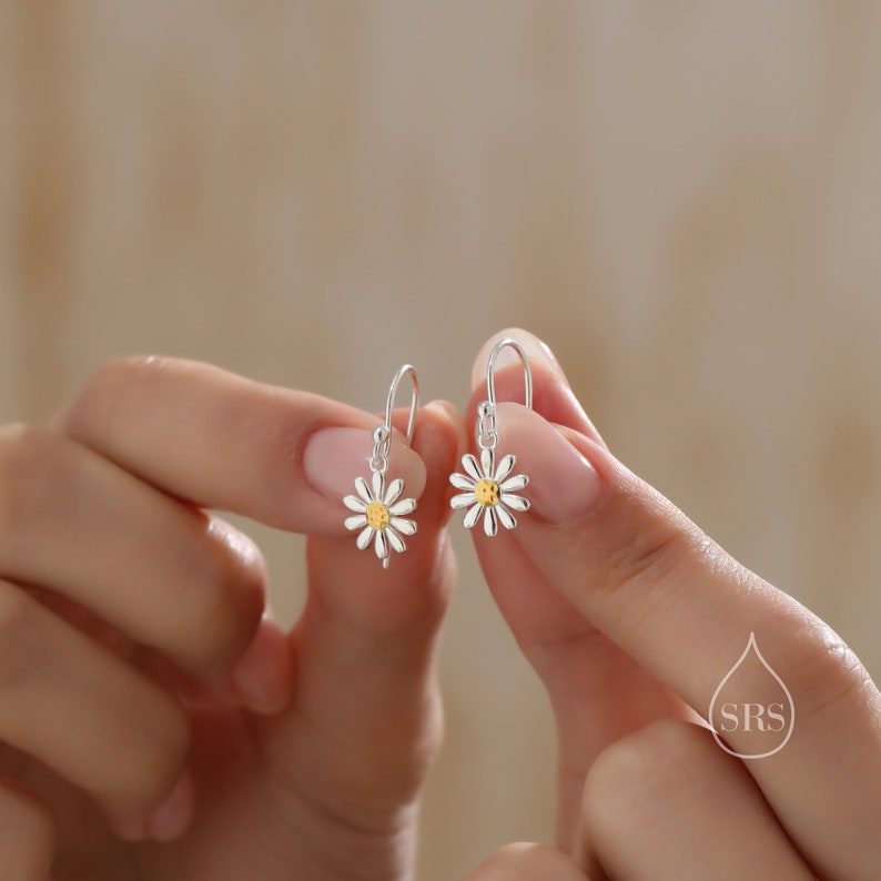 May include: Silver dangle earrings with a small daisy design. The center of the daisy is gold and has a smiley face.