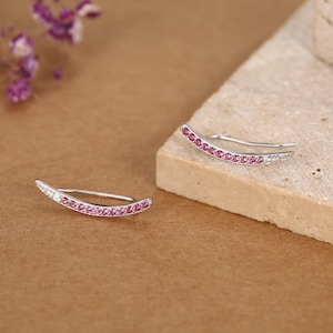 May include: A pair of silver ear climber earrings featuring a curved design set with a row of small, round, vibrant pink gemstones. Each earring has a thin metal post and a sleek, polished finish, showcasing a delicate and symmetrical appearance.
