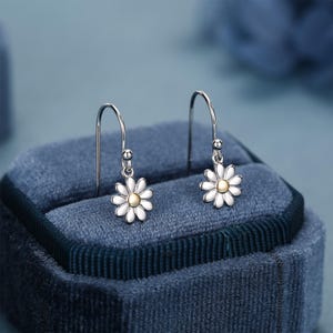 May include: A pair of daisy drop earrings. Each earring features a white daisy flower with a gold-colored center. The earrings are suspended from silver-colored hooks. The earrings are displayed on a blue velvet surface.