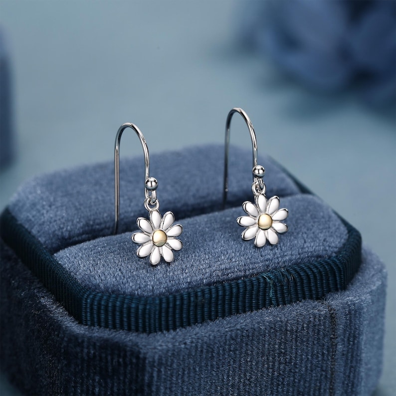 May include: A pair of daisy drop earrings. Each earring features a white daisy flower with a gold-colored center. The earrings are suspended from silver-colored hooks. The earrings are displayed on a blue velvet surface.