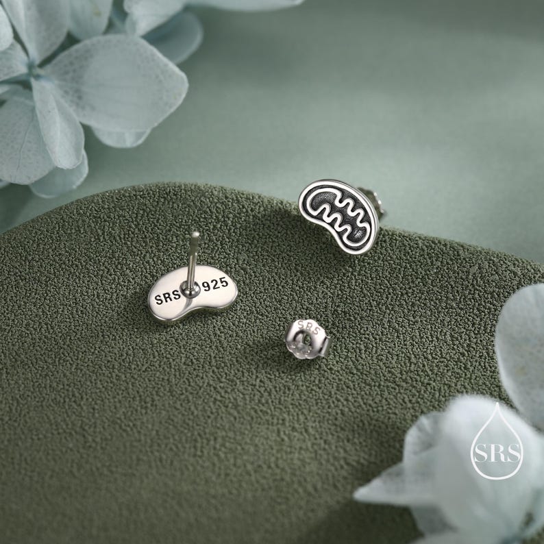 May include: Silver-toned stud earrings with a unique design, featuring a stylized shape with a black outline. The earring post is visible, with the text "SRS 925". The earring back is also visible. The earrings are on a textured green surface.