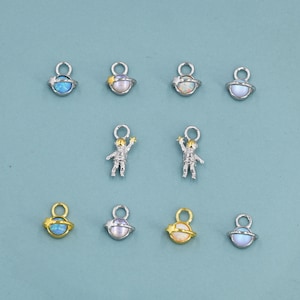 May include: A collection of silver-toned and gold-toned charms. The charms include astronaut figures and planet designs with opalescent and colorful accents. The charms are small and detailed, suitable for jewelry making or crafting projects.