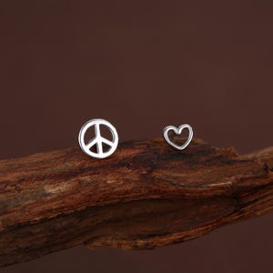 May include: A pair of silver stud earrings. One earring is a peace symbol, the other a heart shape. Both are small and shiny. The peace sign is a well-known emblem, and the heart adds a romantic touch. These earrings are suitable for daily wear.