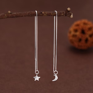以下が含まれることがあります： シルバーのスレッダーピアス。片方のピアスには星のチャーム、もう片方には三日月のチャームが付いています。繊細なチェーンからぶら下がるピアスは、ミニマルで天体的なデザインです。茶色の背景にピアスが表示されています。