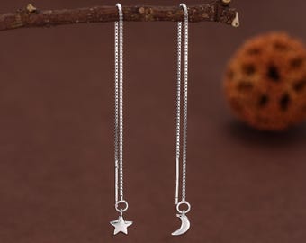 Può includere: Un paio di orecchini a filo d'argento. Un orecchino presenta un ciondolo a forma di stella, l'altro una luna crescente. Gli orecchini pendono da delicate catene, creando un design minimalista e celeste. Gli orecchini sono su sfondo marrone.