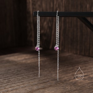 May include: A pair of silver chain earrings with pink gemstone accents. The earrings are hanging from a black surface.