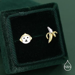 Può includere: Orecchini a bottone a forma di faccia di scimmia e banana. L'orecchino a scimmia ha una faccia bianca con dettagli neri e bordo dorato. L'orecchino a banana è dorato con punta argentata. Presentati in un cofanetto di velluto verde scuro.