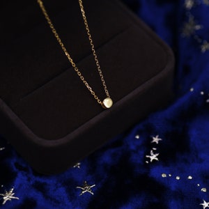 May include: A delicate gold chain necklace with a small, round pendant. The necklace is displayed in a black velvet box on a blue velvet background.