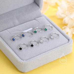 Op de afbeelding: Een set van zes zilveren oorringen met kleine, kleurrijke edelstenen. De oorringen worden getoond in een grijze fluwelen doos. De edelstenen zijn zwart, wit, blauw en groen.