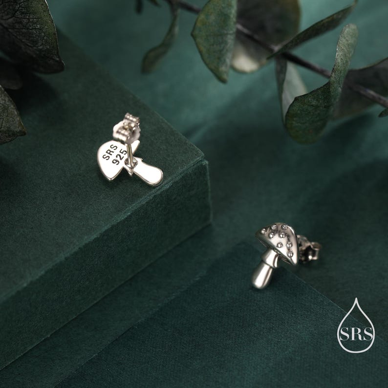 May include: A pair of silver mushroom stud earrings. One earring is in focus, showing the post and the text "SRS 925". The other earring is in the background. The earrings are on a dark green surface with leafy accents.