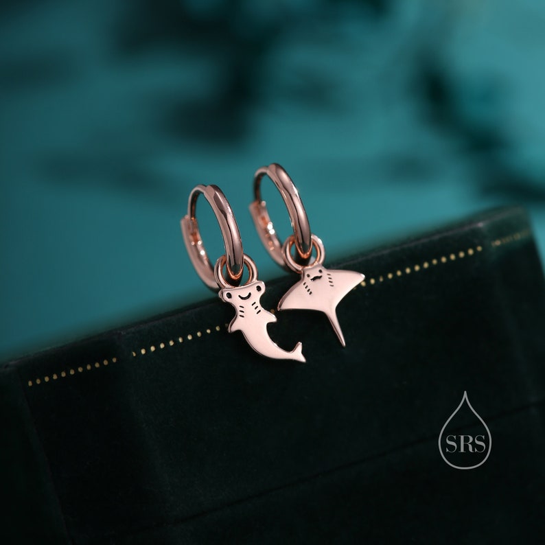May include: A pair of rose gold hoop earrings with a stingray and a hammerhead shark charm. The earrings are on a green velvet surface.