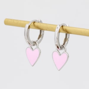 May include: A pair of silver hoop earrings with pink heart charms. The earrings are hanging from a wooden display stand.
