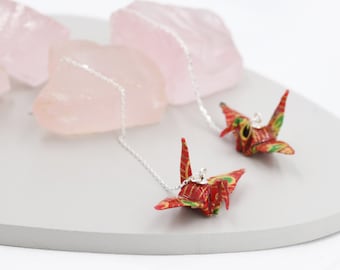 May include: A pair of red and gold origami crane earrings with silver chains. The earrings are hanging from pink crystal rocks.