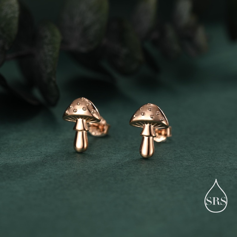 May include: A pair of gold-colored mushroom stud earrings. Each earring features a mushroom cap with small, evenly spaced dots and a stem. The earrings are set against a dark green background.