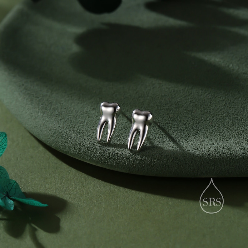 May include: A pair of silver-toned stud earrings shaped like human teeth with two distinct roots. The earrings have a polished, reflective metallic finish and include a small logo in the corner featuring a water drop icon and the letters SRS.