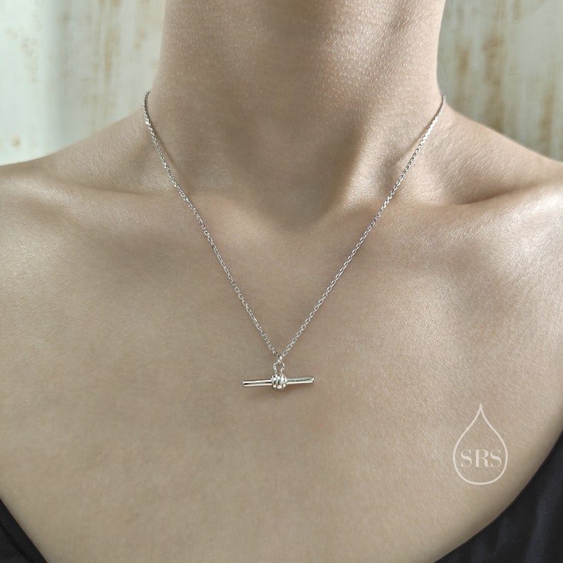 May include: A silver chain necklace with a toggle clasp pendant. The pendant is a simple, straight bar with a round knot at the center.