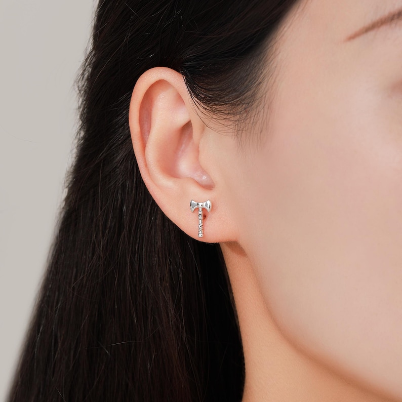 May include: A silver-colored axe-shaped stud earring. The earring is small and detailed, with a miniature axe head and handle. The earring is worn on the earlobe.