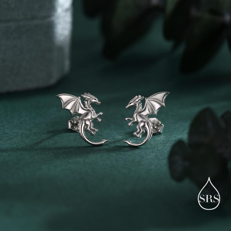 May include: A pair of silver dragon stud earrings. Each earring depicts a detailed dragon with outstretched wings and a curved tail. The earrings are presented on a dark green surface, with a blurred background.