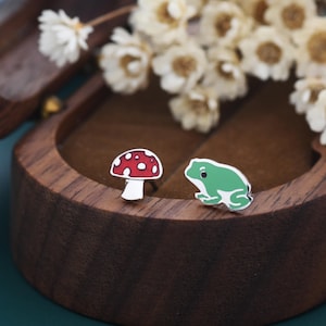 May include: A pair of quirky stud earrings. One earring is a red mushroom with white spots and a white stem. The other is a green frog. The earrings are displayed in a wooden jewellery box.