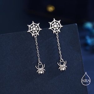 Op de afbeelding: Zilverkleurige oorhangers met een spinnenwebontwerp aan de bovenkant, verbonden door een ketting met een spinnenbedel. De oorbellen worden getoond tegen een donkerblauwe achtergrond.
